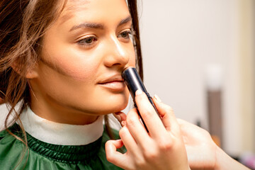 Beautiful young brunette woman receiving makeup with stick concealer on her face in a beauty salon