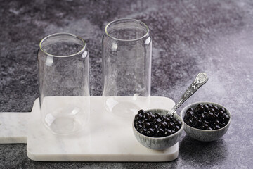 Cooked tapioca pearls for trendy bubble boba ice tea in two small grey ceramic bowls and two transparent empty glasses on marble board on dark grey concrete background