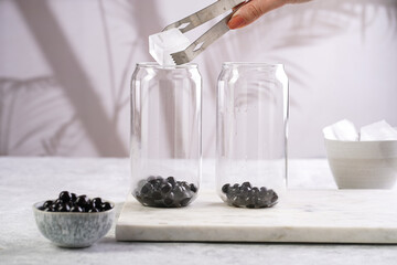 Female hand putting ice into a transparent glass with cooked tapioca pearls for trendy bubble boba ice tea in two small grey ceramic bowls on marble board on dark grey concrete background