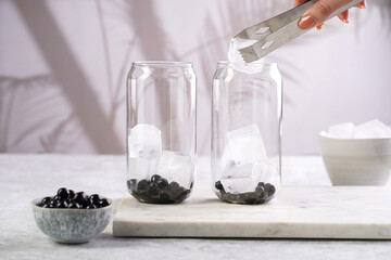 Female hand putting ice into a transparent glass with cooked tapioca pearls for trendy bubble boba ice tea in two small grey ceramic bowls on marble board on dark grey concrete background