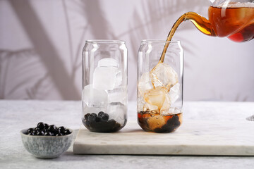 Female hand pouring black tea into transparent glass with ice cubes and cooked tapioca pearls for trendy bubble boba ice tea, two small grey ceramic bowls on marble board on grey concrete background