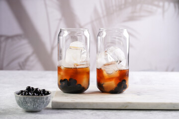 black tea in transparent glass with ice cubes and cooked tapioca pearls for trendy bubble boba ice tea, two small grey ceramic bowls on marble board on grey concrete background