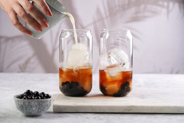 Female hand pouring milk into glass with black tea, ice cubes and cooked tapioca pearls for trendy bubble boba ice tea, two small grey ceramic bowls on marble board on grey concrete background