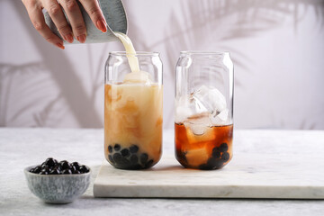 Female hand pouring milk into glass with black tea, ice cubes and cooked tapioca pearls for trendy bubble boba ice tea, two small grey ceramic bowls on marble board on grey concrete background