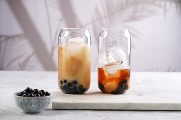 Two glasses with black tea, milk, ice cubes and cooked tapioca pearls for trendy bubble boba ice tea, two small grey ceramic bowls on marble board on pastel tropical background