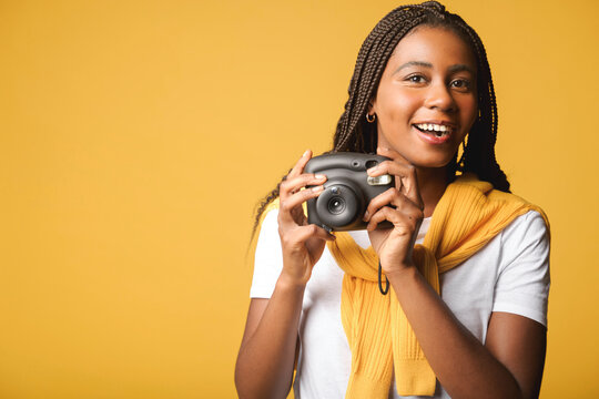 Say cheese. Charming cheerful african-american woman holding polaroid camera standing isolated on yellow, tourist woman making instant photo with retro camera