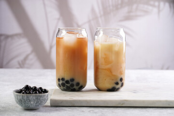 Fototapeta na wymiar Two glasses with black tea, milk, ice cubes and cooked tapioca pearls for trendy bubble boba ice tea, two small grey ceramic bowls on marble board on pastel tropical background