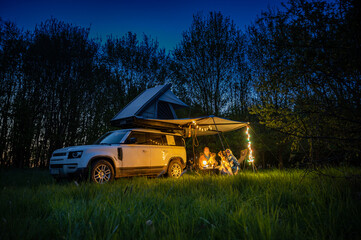 Man and women with dogs camping in front of a 4x4 Offroad vehicle with roof tent at night time and romantic lighting