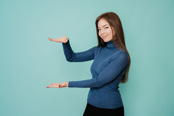 Cheerful young woman with long loose hair in blue sweater, black pants holds an imaginary object with her hands, presents it, looks at the camera, smile on turquoise background in the studio. Mockup.