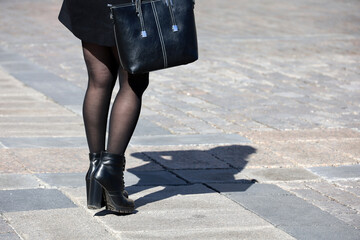 Female legs in black stockings and shoes on high heels. Girl in coat standing on a street with handbag, fashion in autumn city