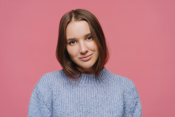 Fototapeta premium Close up shot of satisfied dark haired young lady tilts head, enjoys lovely casual talk, wears warm winter sweater, poses alone against pink background, gazes focused at camera, feels relaxed