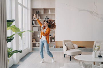 Energetic woman holding smartphone, having fun, dances to music rejoicing with good news at home