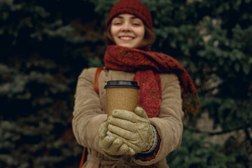 Cheerful woman warming hands with coffee in takeaway blank paper cup in cold weather in winter park 