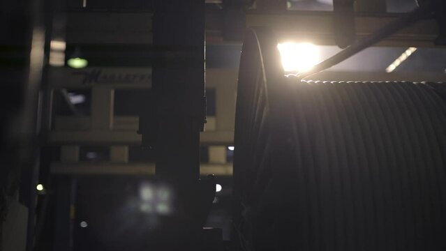 The cable is slowly wound onto a large metal spool. Cable factory. Close-up. Mechanical production process. A large lamp on the ceiling of the factory shines brightly into the camera. 4K