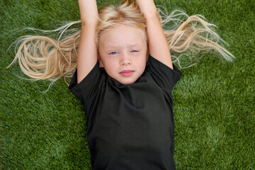 Close up portrait pretty blonde girl lying and stretching on green grass. Mock up black t shirt. Top view.