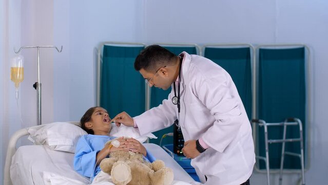 A male Indian doctor with a tongue depressor inspects a little girl's throat for infection - medical examination  pediatrician. A sweet kid admitted to hospital for illness - health issues  sick ch...
