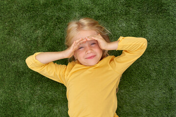 Portrait cute blonde girl eyes lying on green grass. Mock up yellow t shirt. Top view.