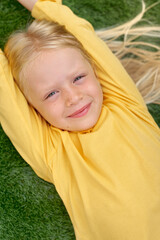 Portrait cute blonde girl eyes lying, stretching on green grass. Mock up yellow t shirt. Top view.