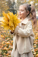 smiling happy little child girl in coat and dress holding autumn leaves, having fun in fall forest