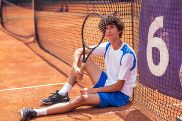 In front of the camera handsome guy sitting down on the outside tennis court he looking to the camera and holding his racket smiling cute