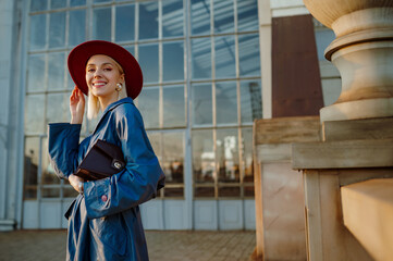 Fashionable happy smiling woman wearing marsala color hat, pearl earrings, blue trench coat,...