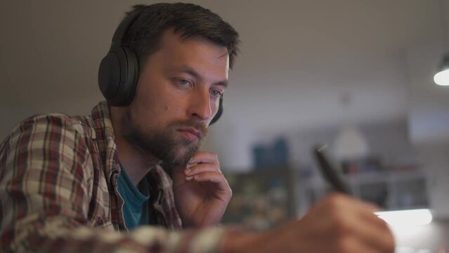 Male American student studies online at home in studio apartment sitting at desk with laptop and taking notes in workbook. Student uses earphones to write notes, watches video online webinar. 