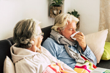 Two person man and woman old age with influenza flu symptoms at home healing and helping each other. Senior couple with cold and virus contagion sitting on the sofa. Cold home temperature for energy - obrazy, fototapety, plakaty