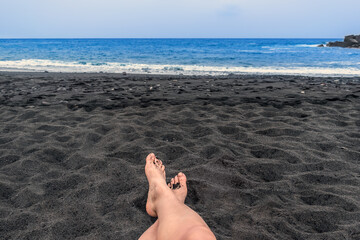 Girl resting on a black sand beach in the Canary Islands, Spain.  Female legs among the volcanic sand against the background of the blue water of the Atlantic Ocean in Tenerife - Powered by Adobe