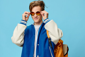 a handsome, stylish guy student with a backpack on his back in a blue bomber jacket puts on his bright sunglasses