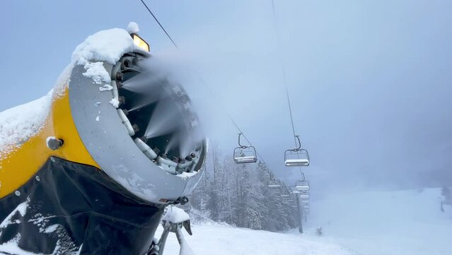 4K yellow snow-making gun producing snow on ski slope with cableway in background. Skiing, equipment, skiing resort concept.