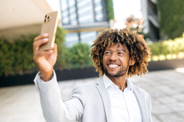 Selfie photo concept. Positive handsome curly haired mixed race man standing outdoors, taking a selfie on her smartphone, mixed race male blogger having live streaming on social network, smiling