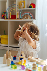 Toddler looking through the colorful window of a toy. Educational game for baby in modern nursery. A little girl looking through transparent colored block toy. Wooden rainbow stacking blocks.