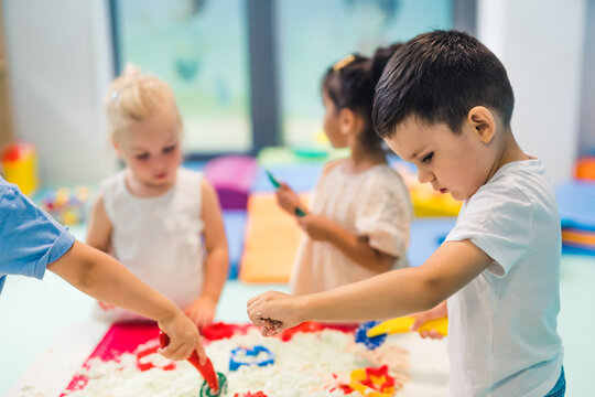 Relaxing sensory play with moldable kinetic sand at nursery school. Toddlers with their teacher having fun around the table using different tools for sculpting sand such as colorful and textured