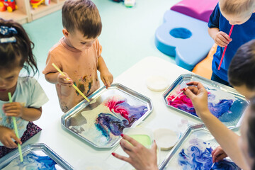 Multi-cultural nursery school. Toddlers playing with striped straws and milk painting, using nontoxic food coloring for colors. Creative kids activity for using their senses and brain development - obrazy, fototapety, plakaty