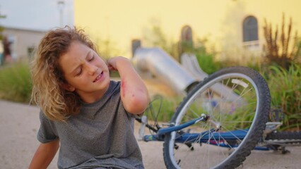 The boy sits on the road after falling off a bicycle, calms the pain in his elbow. Broken bike in the background