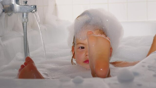 A little cheerful girl washes and plays in the bath with foam. Daily hygiene and skin care of children. Playing with water. Baby shampoo soap