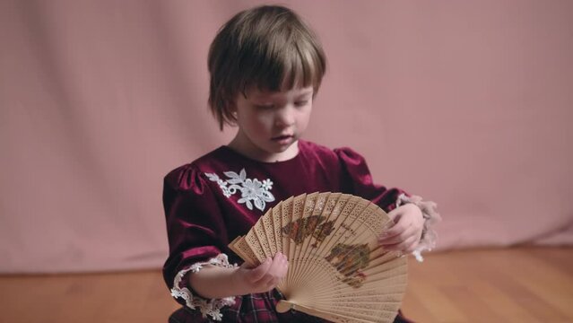 Cute little girl in an vintage burgundy dress is playing with a wooden fan. The concept of children theater productions and developing acting circles. Coral background