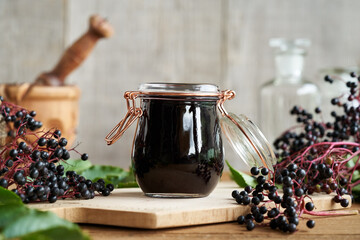 A jar of black elder syrup with fresh elderberries on a table