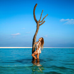 Sexy woman in white bikini on the sun-tanned slim and shapely body is posing on the beach on Maldives island. Perfect  body bikini model in luxury resort on Maldives. - obrazy, fototapety, plakaty