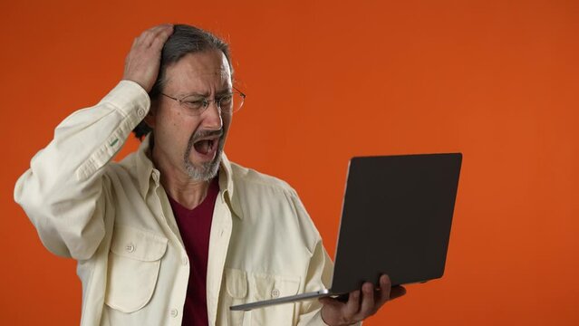 Unhappy bearded mature man 50s casual shirt isolated on solid orange background studio. People lifestyle concept. Working on laptop pc computer showing thumbs down.