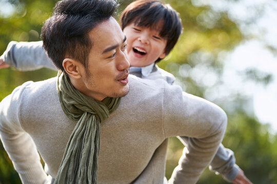young asian father having fun carrying son on back outdoors in park