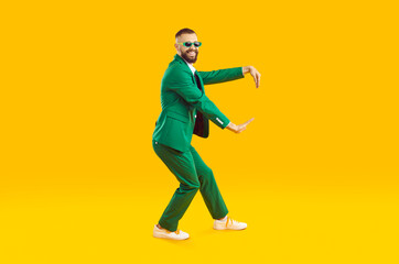 Fototapeta na wymiar Funny young guy dancing in a cool party outfit. Happy, joyful man wearing a stylish green suit and sunglasses dancing isolated on a bright yellow colour background. Full body studio shot, side view