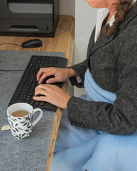 Eine Frau schützt sich mit Decke und heißem Tee am Schreibtisch vor der Kälte bei der Arbeit,...