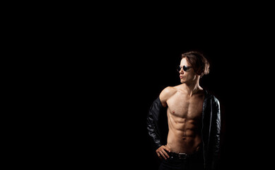 Fototapeta na wymiar Fashionable macho man in sunglasses, naked torso with abs muscles. Long-haired sexy man in a leather jacket on a black background