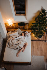 A young woman wrapped in a blanket lies on the bed near the Christmas tree.