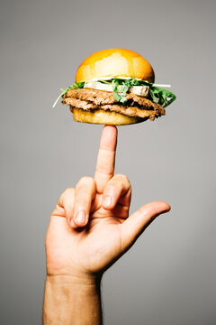 Human Hand Holding A Finger A Doble Burger With Old Dry Cheese And Lettuce Over A Grey Background