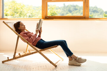 Relaxed teen girl reading paper book, sitting in comfy chair, resting and enjoying free time at home