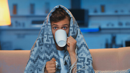 sick man with blue eyes drinking tea while sitting under blanket in living room