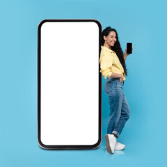 Happy young woman standing near big cellphone with empty white screen, showing mobile device on blue background. Mockup