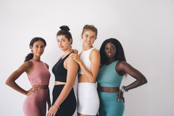 Multiethnic group of healthy curvaceous and slim women in tracksuit posing at white background with copy space in sportive gym studio, half length portrait of diverse fit girls reaching body positive